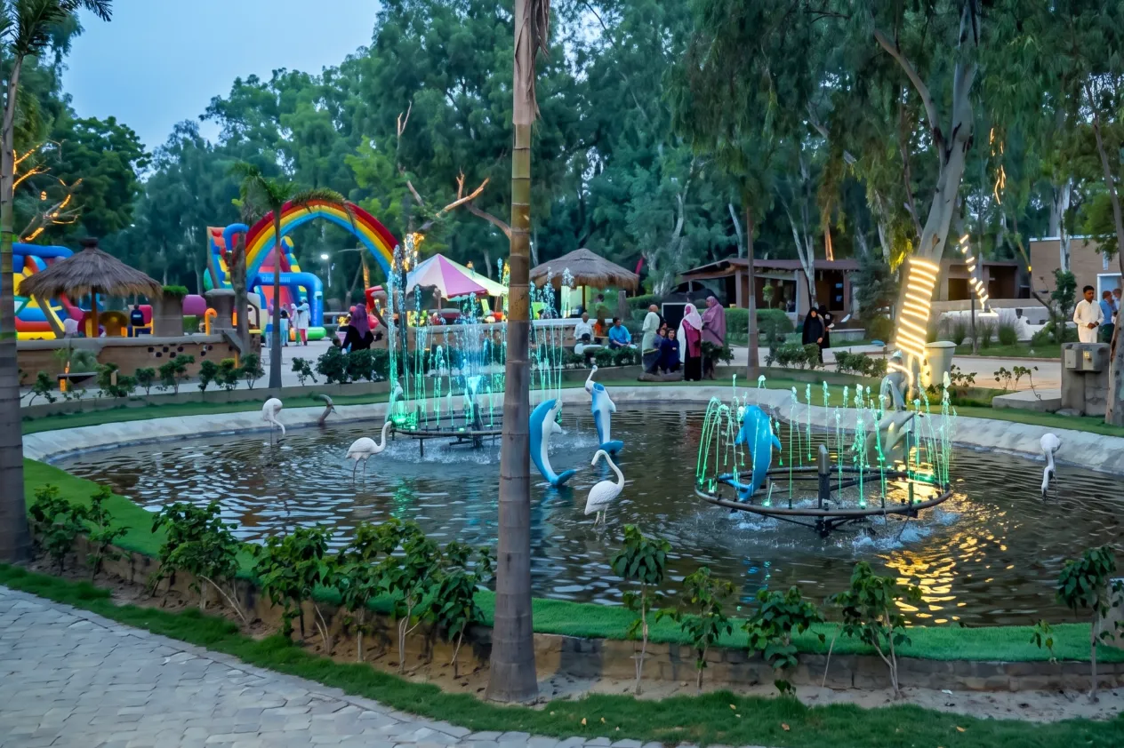 Wide shot of a stone-edged pond featuring glowing water fountains, dolphin and crane statues, surrounded by paved walkways and park greenery.