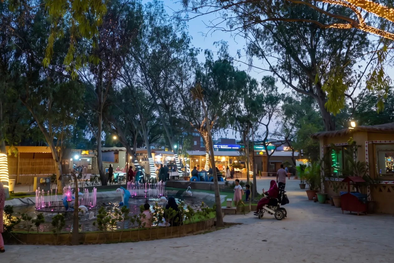 Families strolling past the illuminated pond fountain at dusk under mature trees decorated with warm fairy string lights.