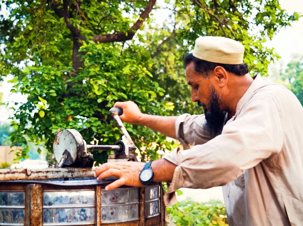Beekeeper in traditional attire operating a manual centrifugal honey extraction machine outdoors at Lahore Organic Village honey farm.